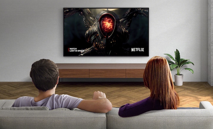 Image of a couple in a living room watching Netflix on a wall-mounted BRAVIA TV. When the Adaptive Calibrated Mode button below the image is toggled on and off, you can see the screen brightness change to match the environment.